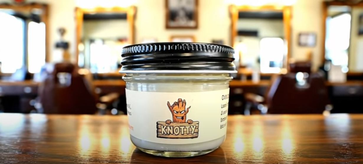 A cylindrical glass jar of Rub N Tug's “Knotty” pomade sits prominently on a polished wooden barber station, centered in the foreground of a traditional barbershop setting. The label features a cheeky cartoon tree character holding a wooden sign that reads “KNOTTY.” The black lid adds contrast, while blurred barber chairs and mirrors fill the bright, warmly lit background, evoking a classic grooming environment. Knotty Rub N Tug styling product, classic grooming aesthetic, handcrafted organic hair care.