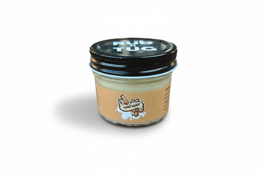 A close-up of the Rub N Tug "Handy Sandy" hair pomade jar, featuring a bold orange label with a playful cartoon log character gripping a banner that reads "Handy Sandy." The black lid prominently displays the Rub N Tug logo in white, and the glass jar rests against a white background.  Handy Sandy by Rub N Tug — a premium, all-natural pomade with cheeky branding and long-lasting hold, developed by barbers for confident, playful styling.