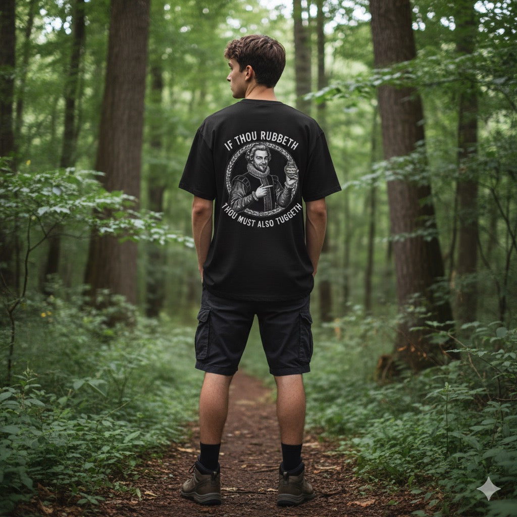 A young man with short hair stands on a forest trail wearing a black Rub N Tug T-shirt that displays a vintage illustration of a gentleman holding a jar of Rub N Tug Pomade and the phrase “If Thou Rubbeth, Thou Must Also Tuggeth.” The scene conveys a modern, outdoorsy lifestyle image for the veteran-owned, all-natural men’s grooming brand.